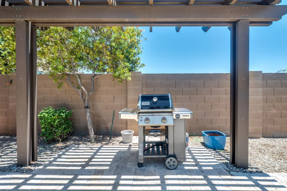 An exterior view of a pergola-shaded patio area complete with a grill, surrounded by greenery and potted plants, creating an intimate spot for outdoor dining and celebrations.