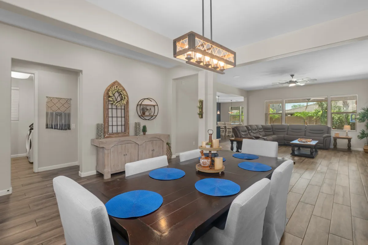 A bright, inviting dining space featuring a formal dining table dressed with blue placemats. The room includes a chandelier, a large decorative wall clock, and comfortable seating, all set near a cozy seating area.