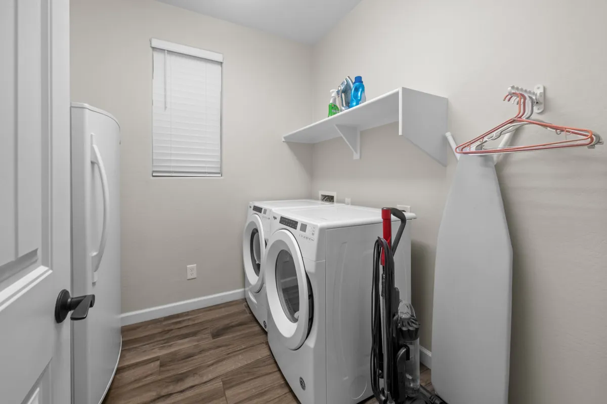A practical laundry room equipped with a washing machine, dryer, shelves stocked with cleaning supplies, an ironing board, and vacuum cleaner. The space is organized and highly functional, catering to daily living needs.