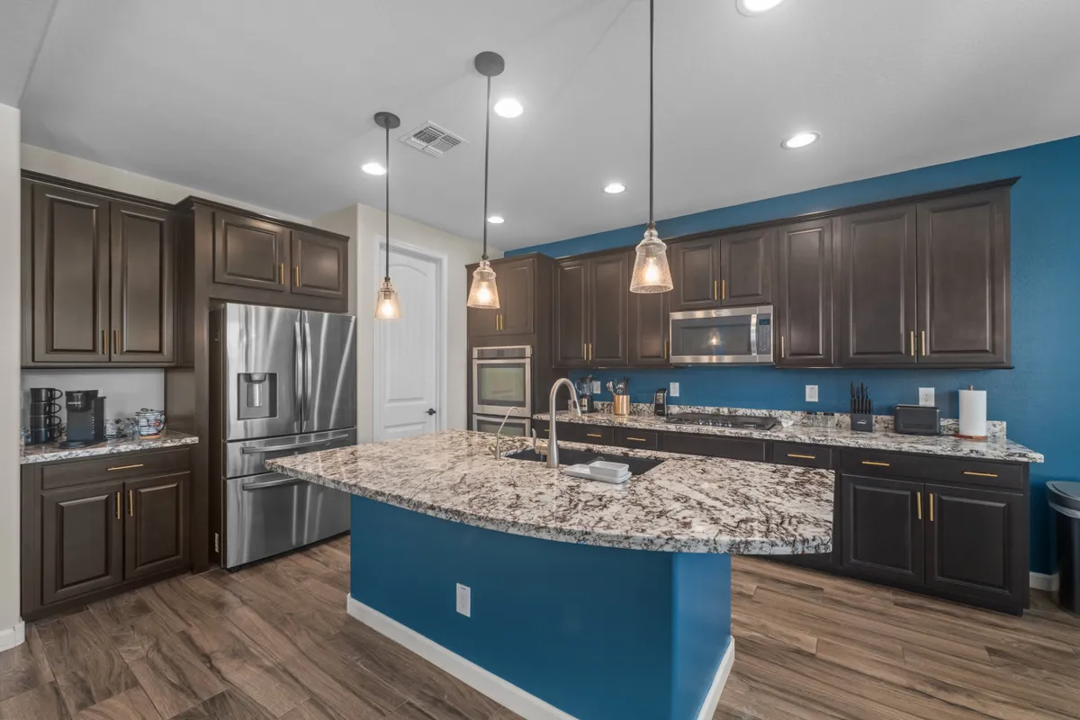 A stylish, modern kitchen featuring a granite countertop island at the center, surrounded by dark cabinets and stainless steel appliances. Pendant lights hang above the island, creating an inviting space for cooking and gathering. The kitchen is well-lit and arranged for both culinary activity and social interaction.