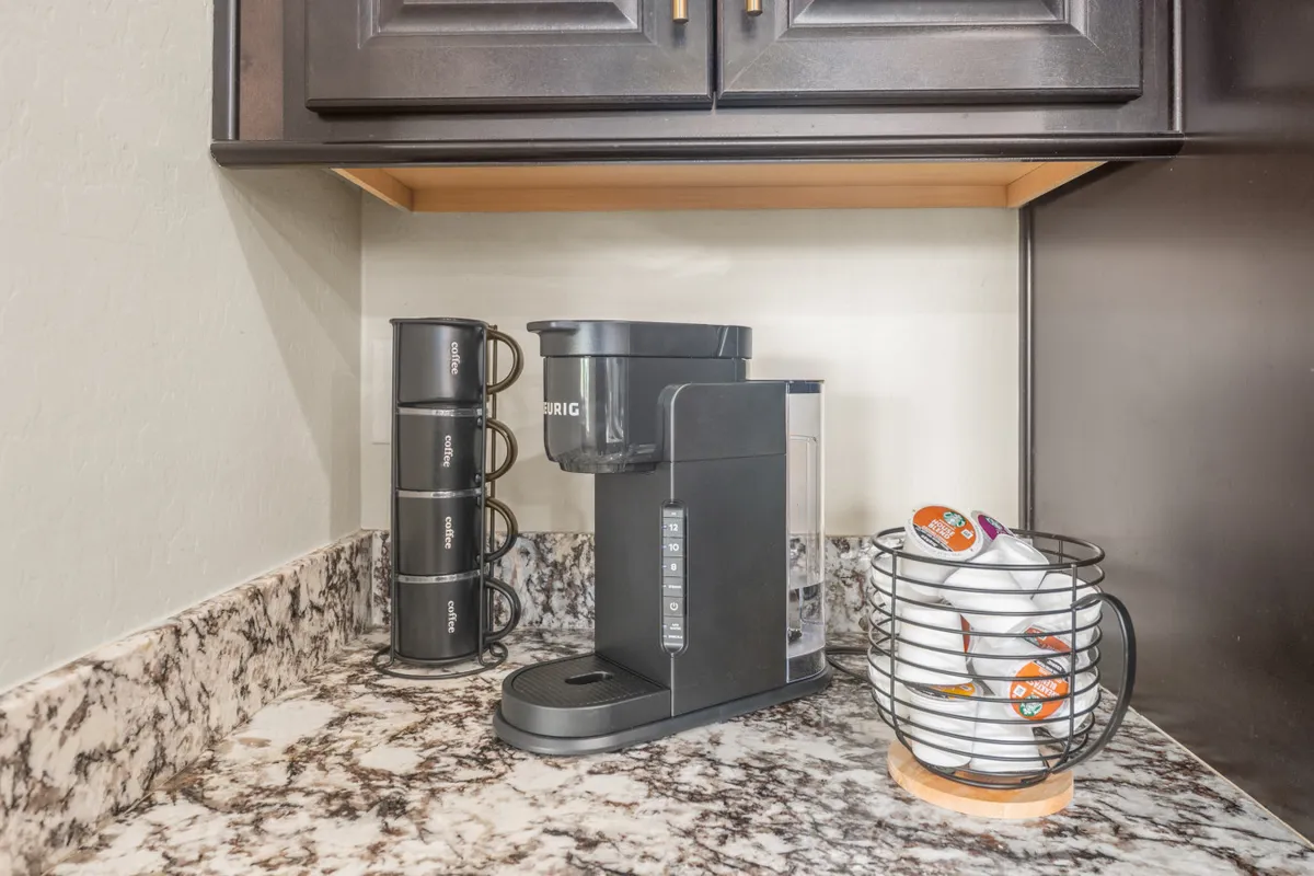 A dedicated coffee bar area, complete with a Keurig coffee maker, a neatly organized pod holder, and a stack of mugs ready for use. The design is welcoming and practical—ideal for quiet mornings or group caffeine breaks.