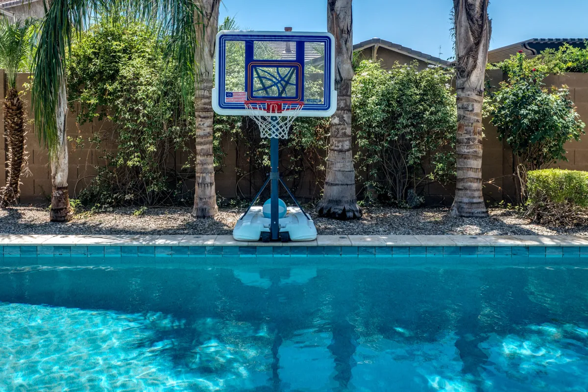 An inviting exterior shot of the swimming pool, featuring a basketball hoop at one end, tall palm trees in the background, and sunbathing areas surrounding the pool. The scene is bright and playful, suggesting an active outdoor environment for both swimming and games.