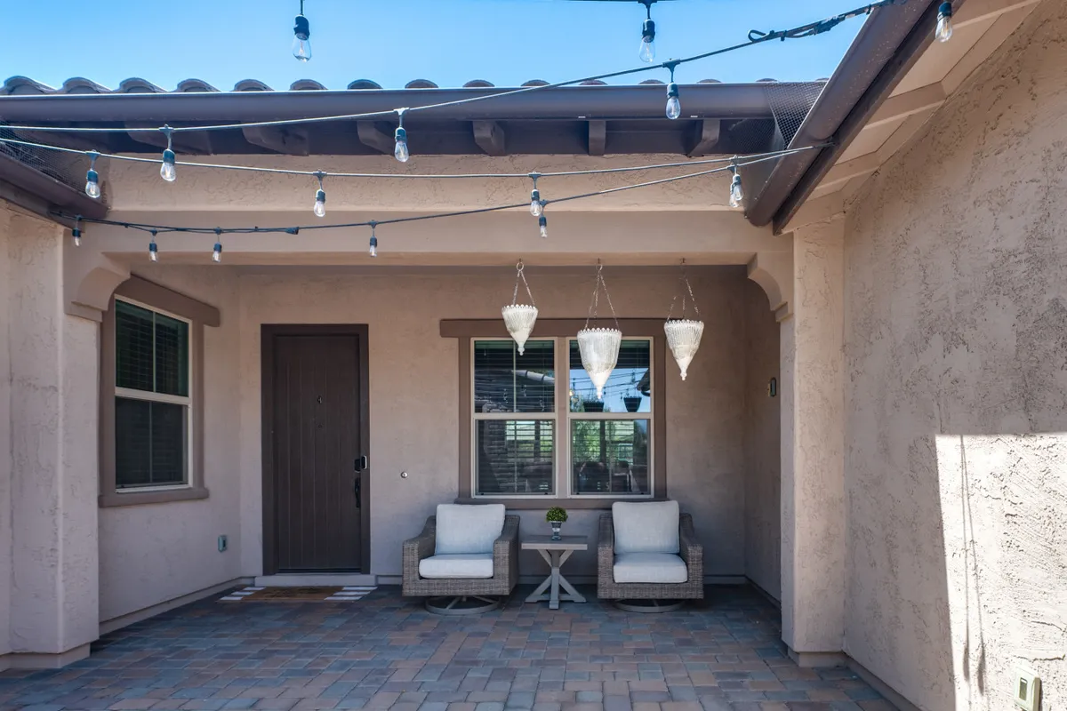 A welcoming front patio with cozy outdoor seating and hanging light fixtures sits beside the home’s front door. The ambiance transitions seamlessly from sunny afternoons to glowing evenings under string lights, suggesting a perfect spot for intimate conversations or a sunset drink.