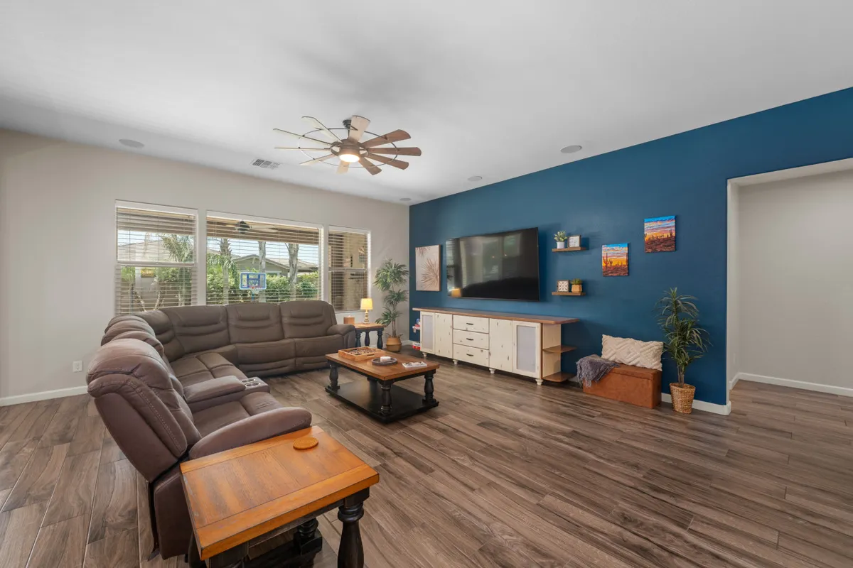 A spacious great room featuring a large, plush sectional sofa facing a sizable TV unit. The coffee table is neatly arranged, and decorative wall art and windows provide natural light and inviting views. The layout is open, offering plenty of space for gathering, relaxing, or playing games.