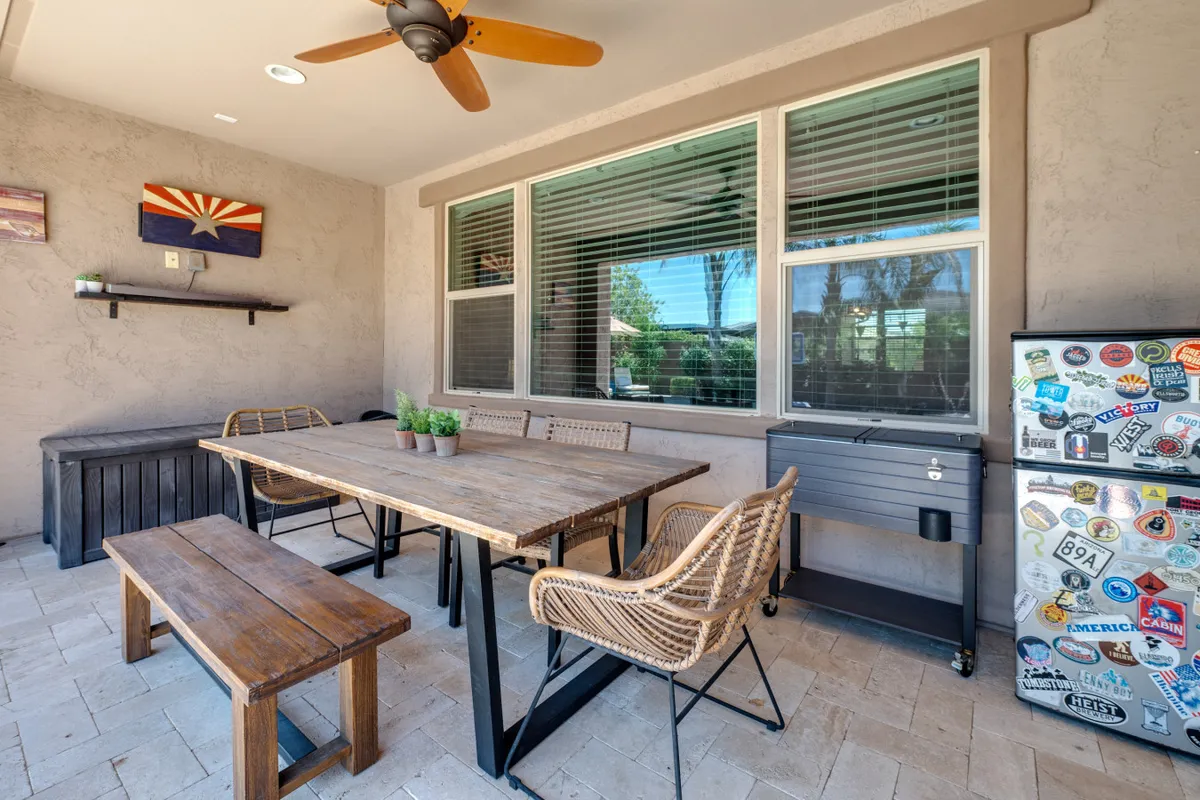 A wooden outdoor dining table with woven chairs sits on the patio, surrounded by decorative wall art and miniature plants. A sticker-covered fridge adds personality, and the setup signals relaxed, social meals in a shaded, comfortable space.