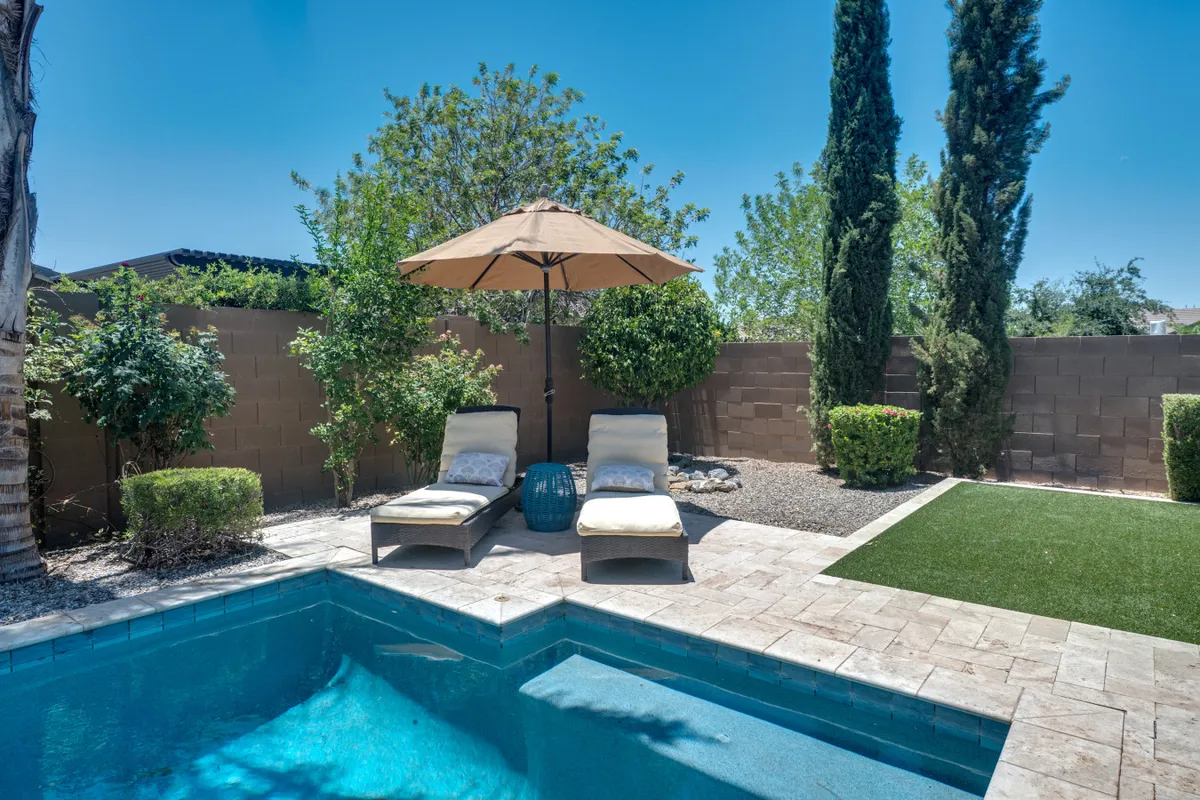 A serene backyard setting with a swimming pool surrounded by lounge chairs, an umbrella for shade, and lush greenery. The pool area looks perfect for relaxation, reading, or unwinding after a day's work.