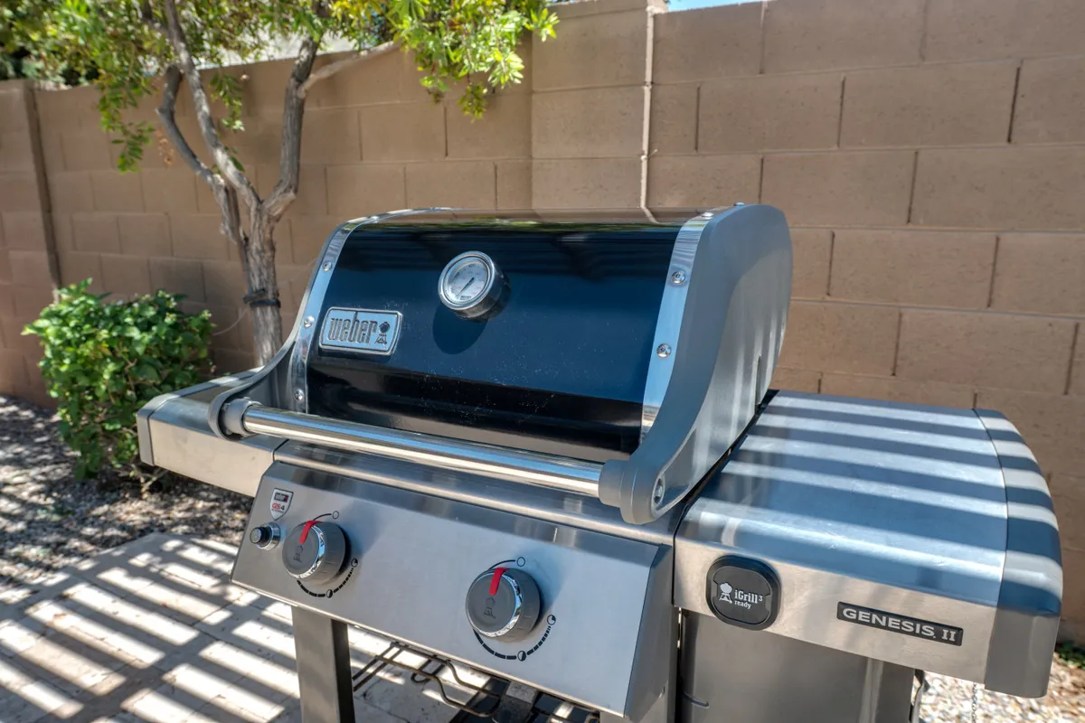The focus is on a Weber propane BBQ grill set against a backdrop of lush greenery and a textured wall. The grill is well-positioned for convenient outdoor cooking, reinforcing the alfresco lifestyle. The ambiance is casual and inviting, perfect for gatherings and meals under the sun or stars.