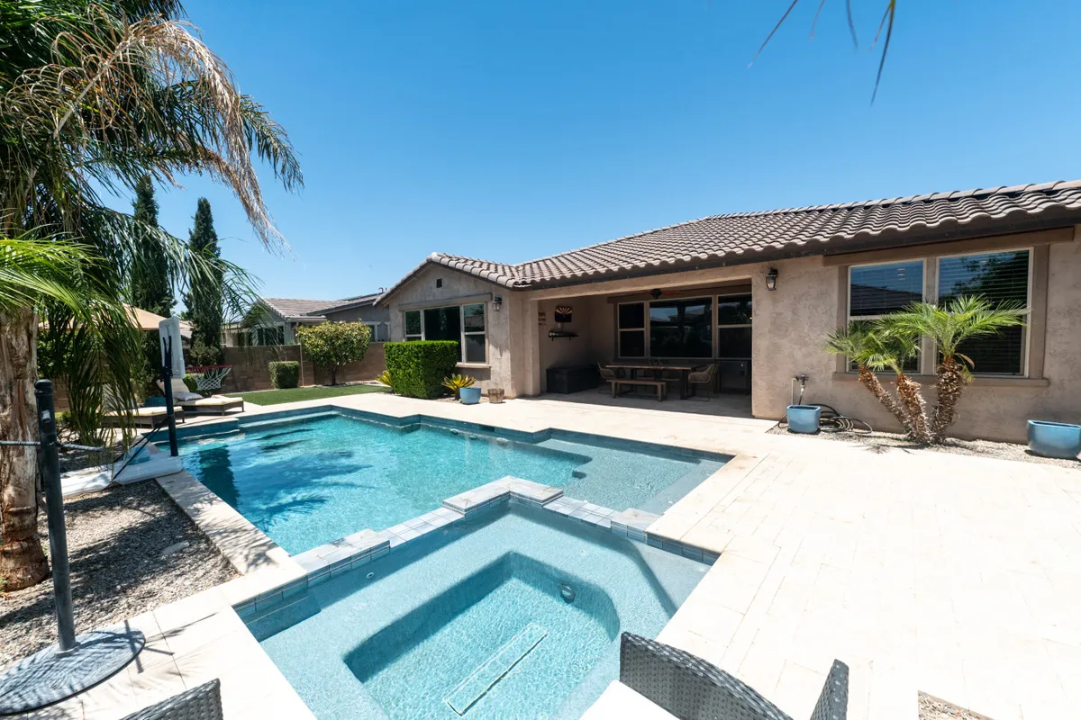 A sun-drenched backyard features a sparkling swimming pool and an inviting jacuzzi, surrounded by palm trees and ample outdoor seating. The house’s exterior creates a sense of privacy and tranquility, while the poolside area is perfect for lounging or sharing morning coffee.