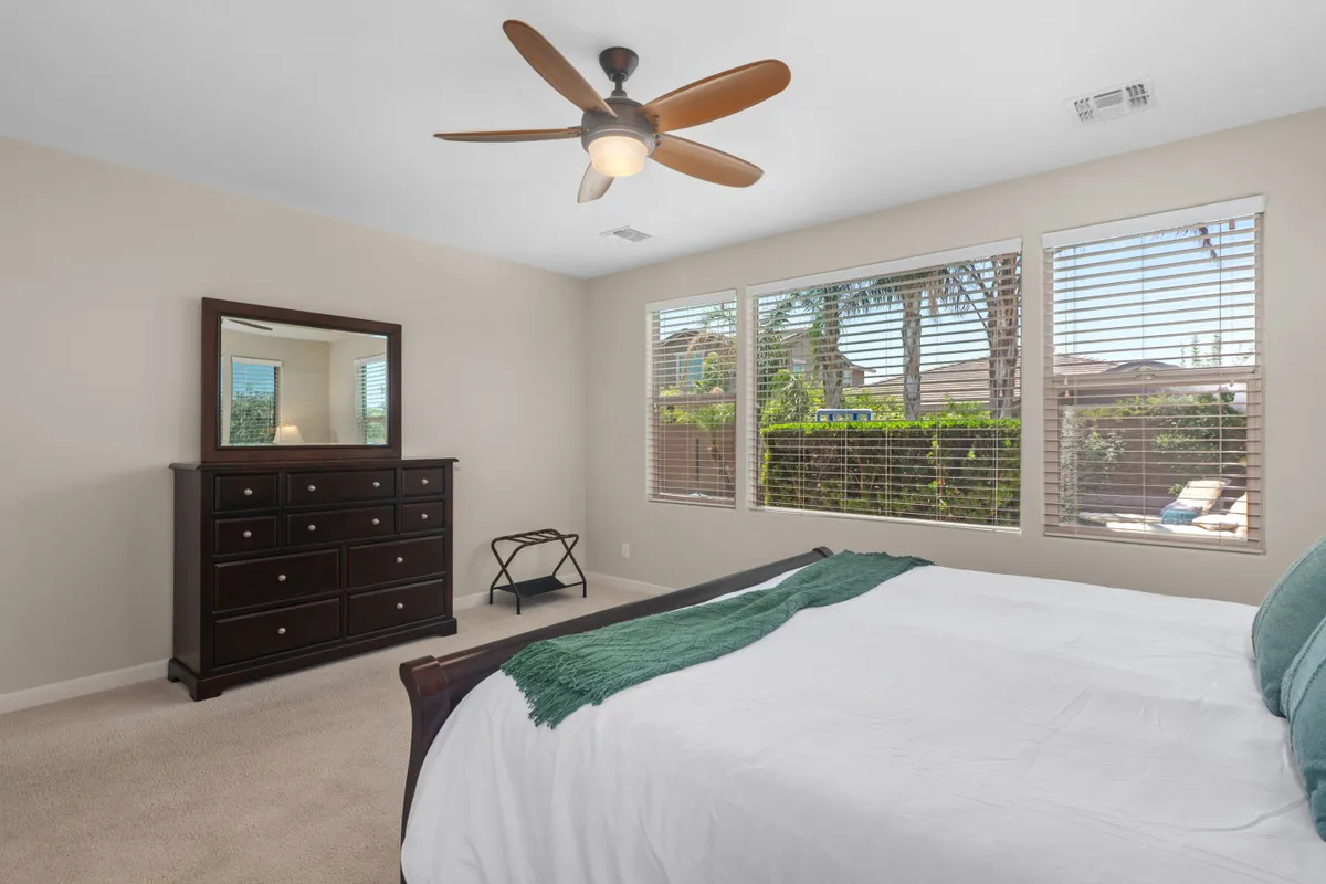A tranquil guest bedroom furnished with a bed dressed in white linens, a dresser with a mirror, and large windows offering lovely views of greenery. The space is filled with natural light, creating a cozy and restful environment.